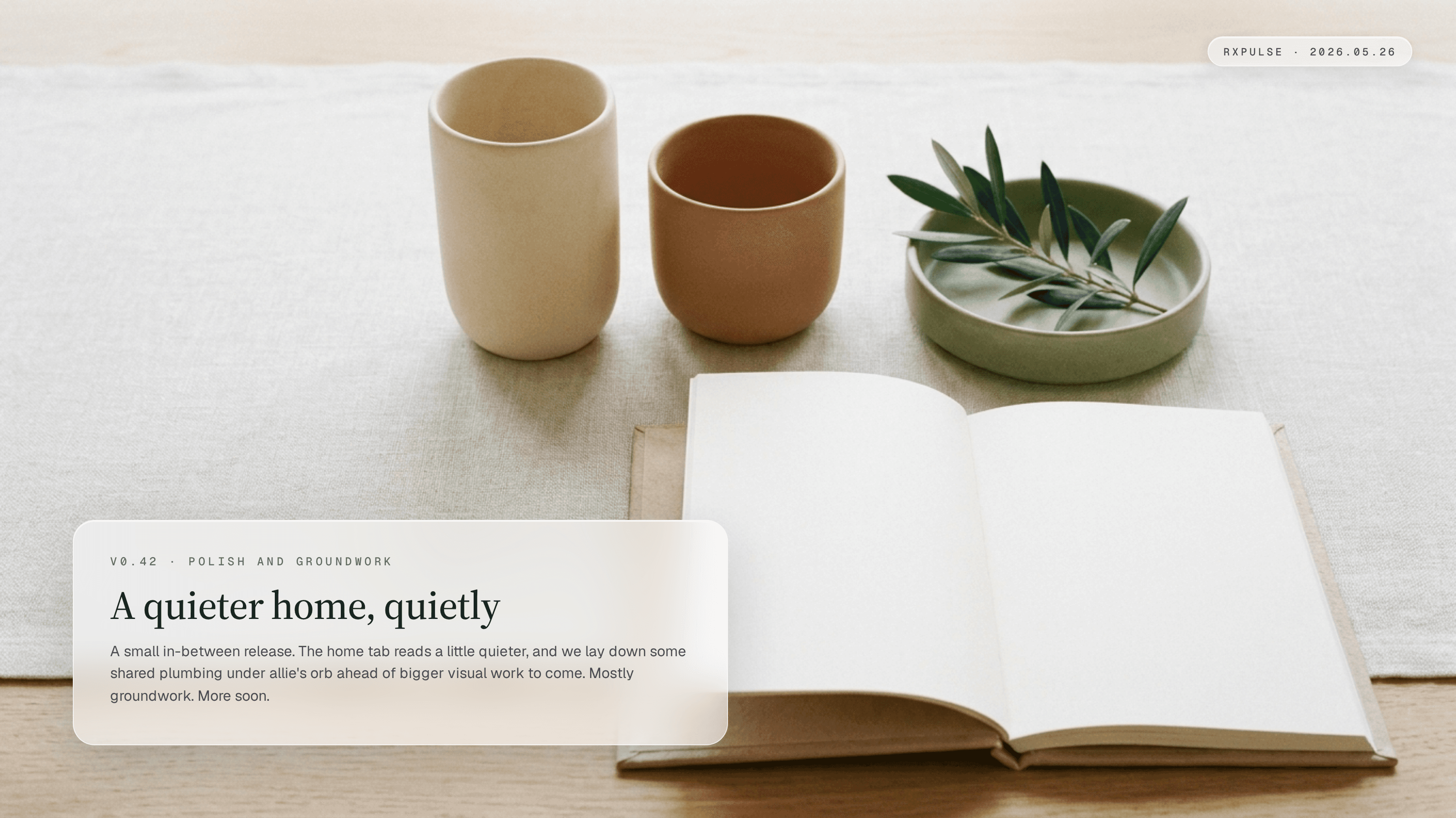 Editorial release artwork. A quiet still life on a warm wooden table: two hand-poured ceramic vessels (cream and terracotta) in close alignment on a clean linen runner, an open journal beside them turned to two facing uncreased pages, a low sage-green ceramic dish holding a small sprig of fresh olive leaves resting at rest, soft midday light from an unseen window. A glassmorphism caption card in the lower-left reads "v0.42 · polish and groundwork" with the headline "A quieter home, quietly" and a short summary: a small in-between release where the home tab reads a little quieter and shared plumbing lands under allie's orb ahead of bigger visual work to come. Mostly groundwork; more soon.