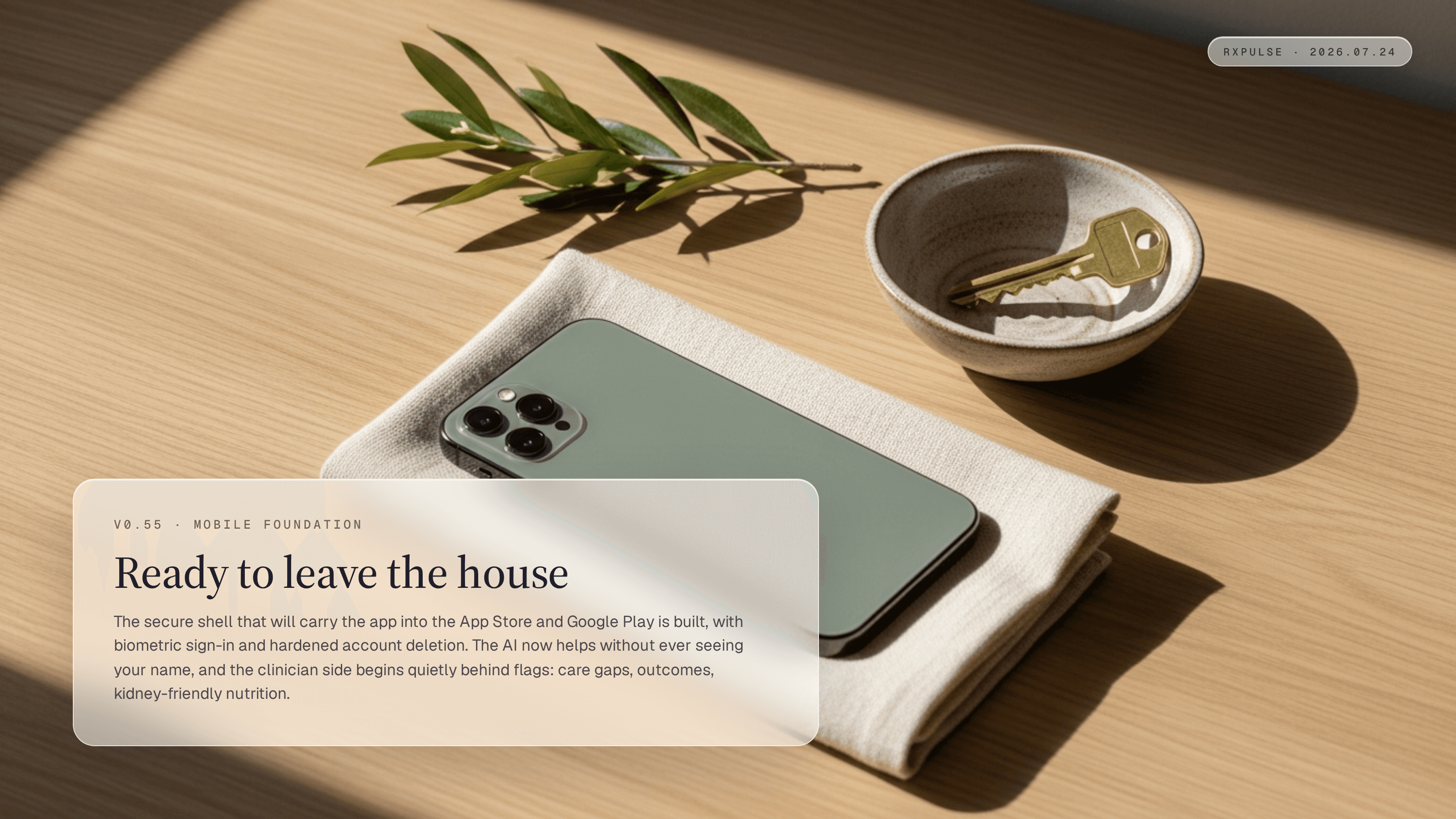 A sage-green phone lying face-down on folded cream linen beside a small ceramic bowl holding a brass house key, with an olive sprig on a pale oak surface in soft window light.